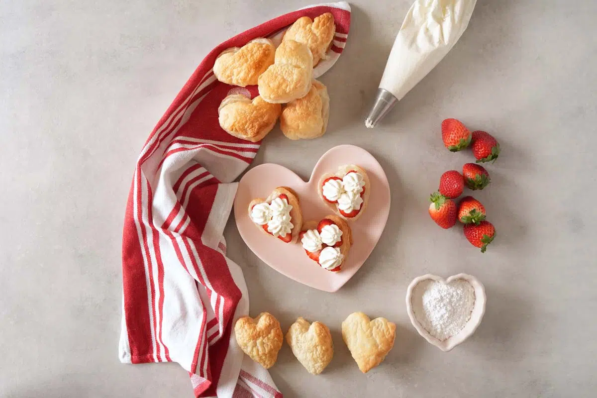 Cream piped onto heart-shaped puff pasty near shells, berries, and a red, striped heart napkin.