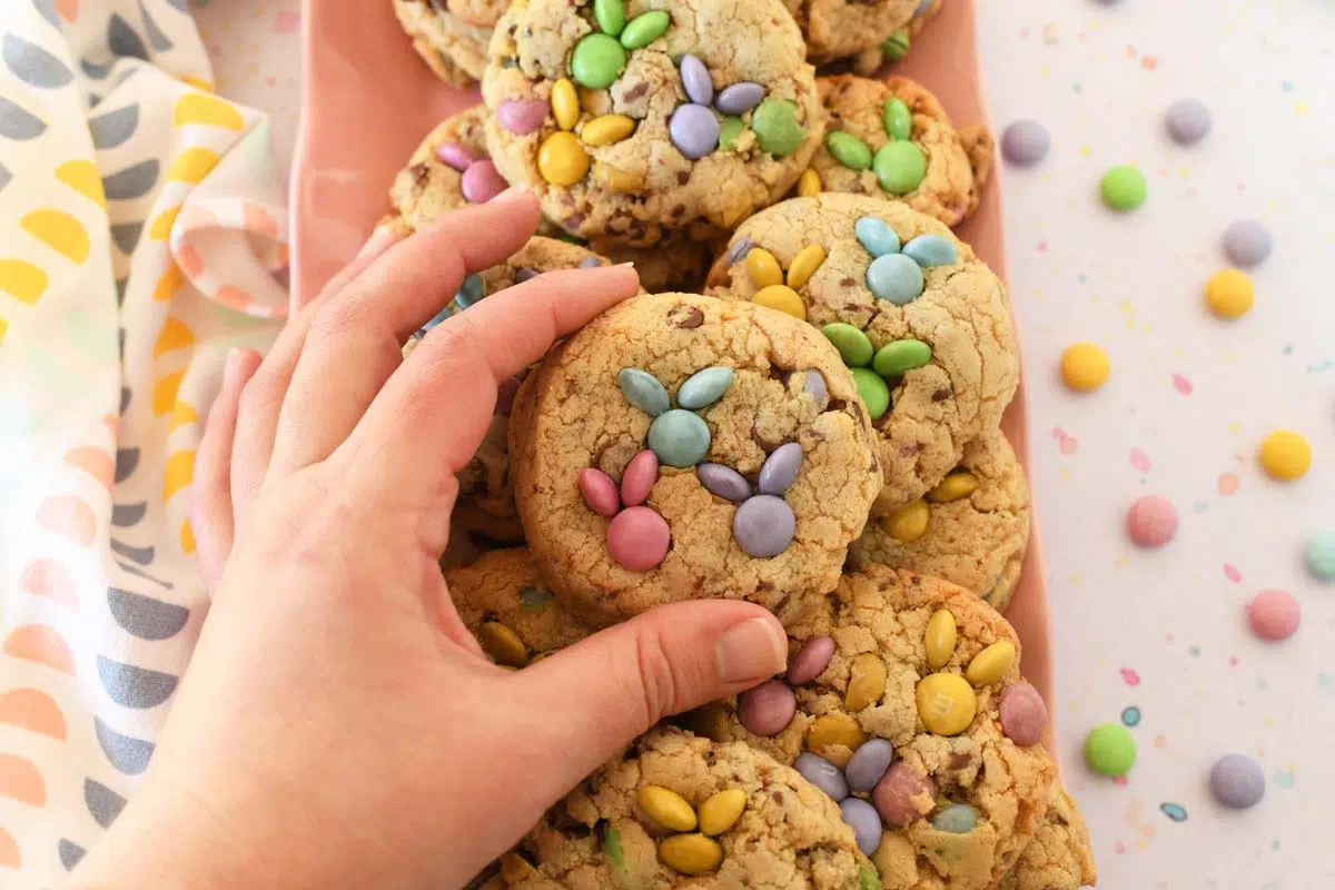 A white hand holding a thick chocolate chip cookie with pastel bunny M&M faces.