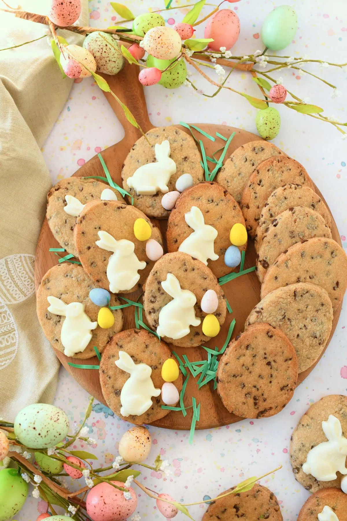 A wooden tray of Chocolate chip egg cookies with white bunnies and Cadbury Eggs near pastel egg florals and a tan napkin.