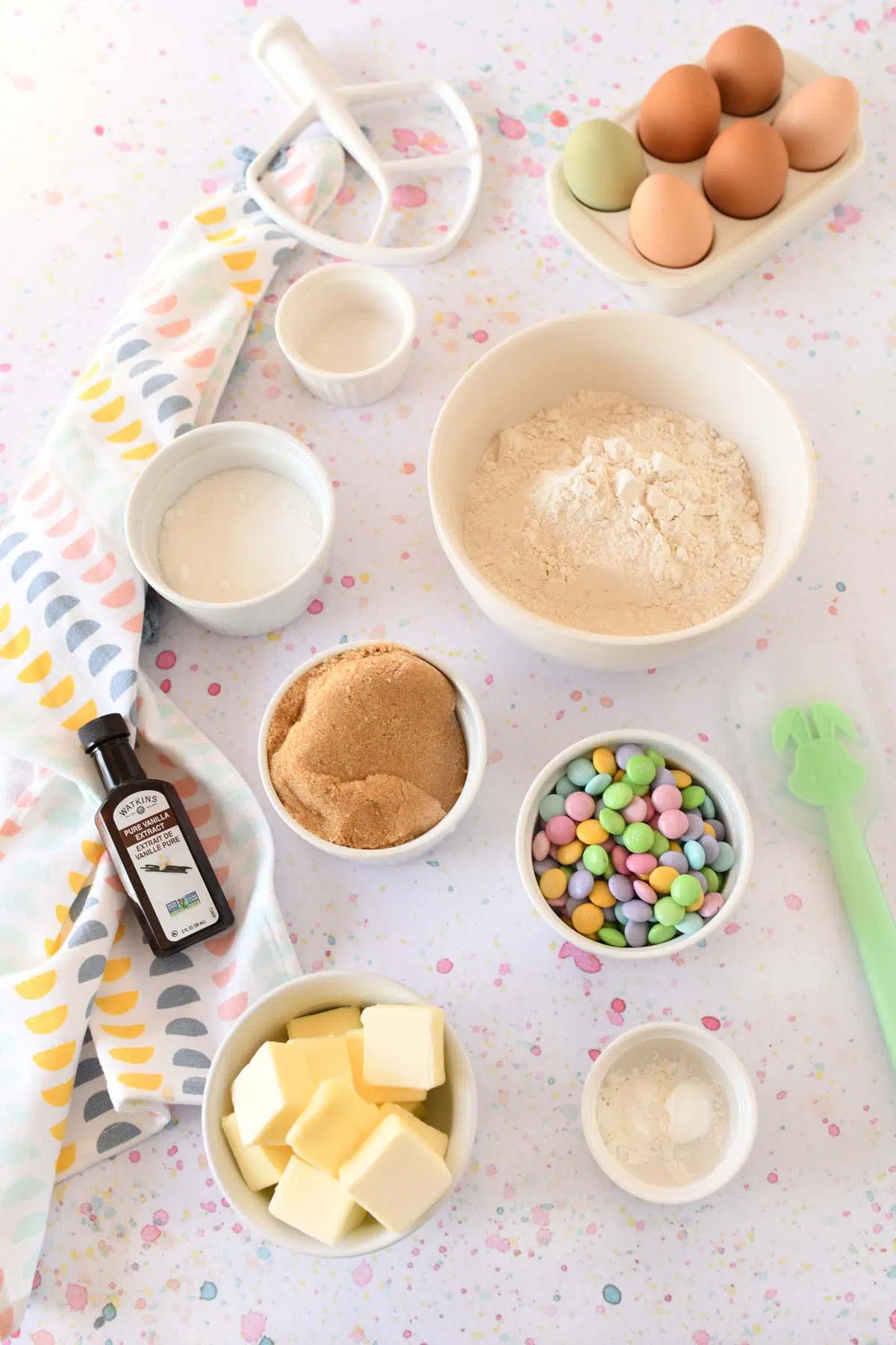 M&M Easter cookie ingredients on a pastel speckled surface. Image also has a pastel printed napkin to the left.
