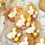 Chocolate chip egg cookies with white bunnies and Cadbury Eggs near green grass clippings and a tan napkin with pastel florals.