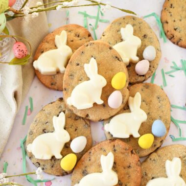 Chocolate chip egg cookies with white bunnies and Cadbury Eggs near green grass clippings and a tan napkin with pastel florals.