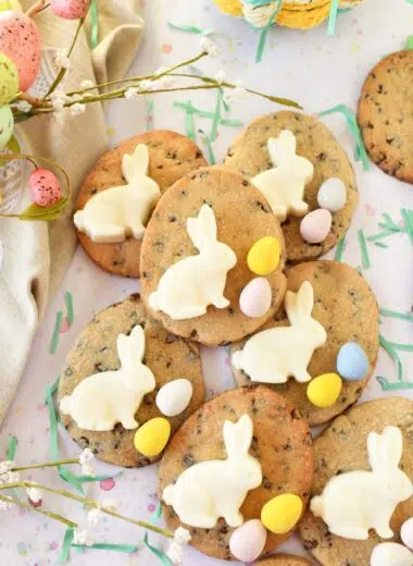 Chocolate chip egg cookies with white bunnies and Cadbury Eggs near green grass clippings and a tan napkin with pastel florals.