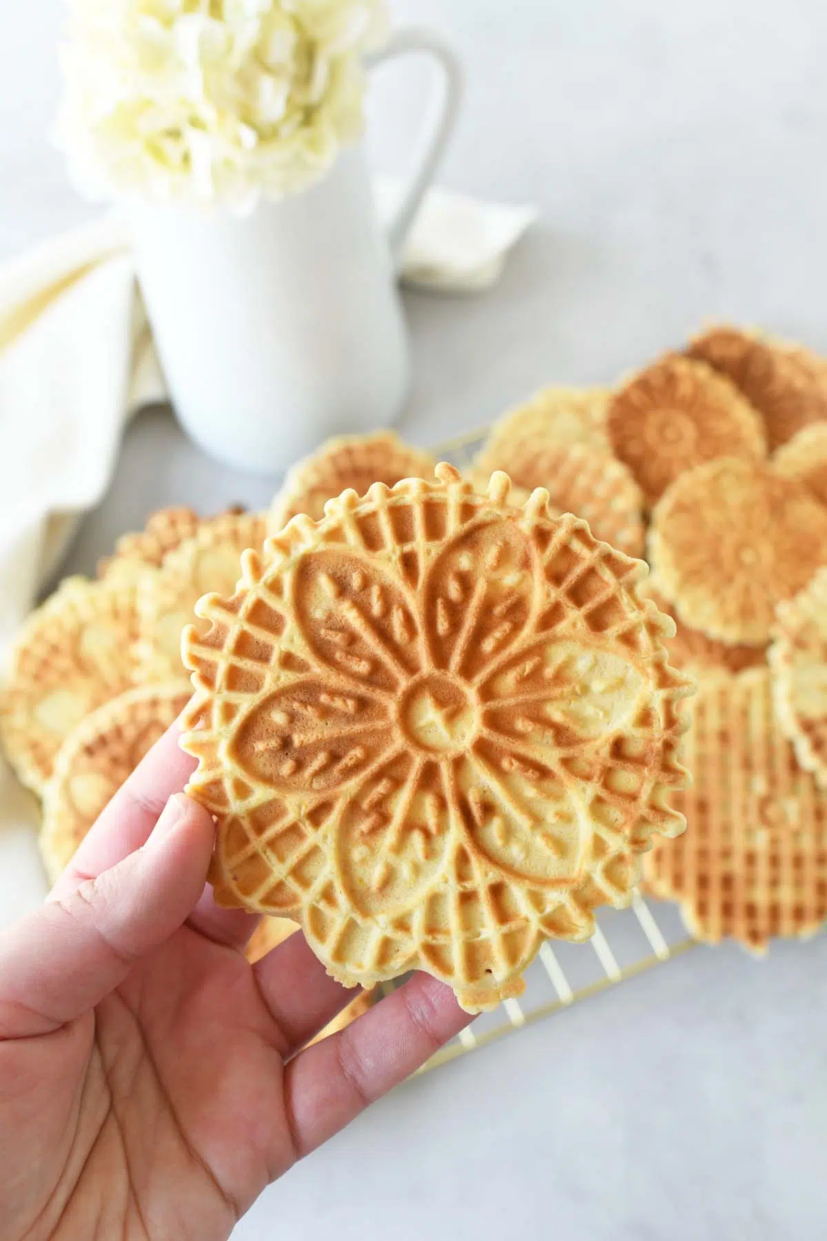 A golden brown pizzelle in a white hand with flowers and a napkin in the background.