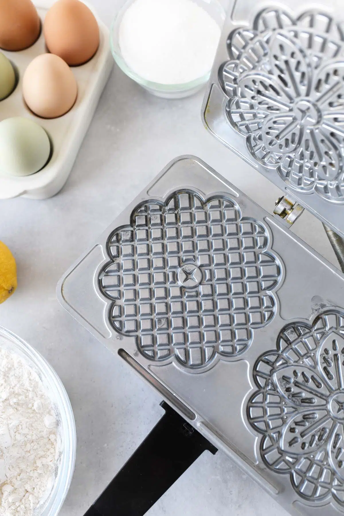 Pizzelle iron near ingredients like colorful eggs on a grey surface.