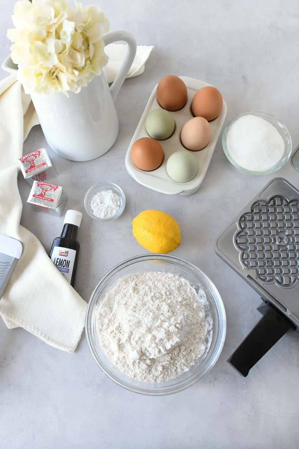 Ingredients to make pizzelles on a grey, stone surface near a white vase with a white flower.