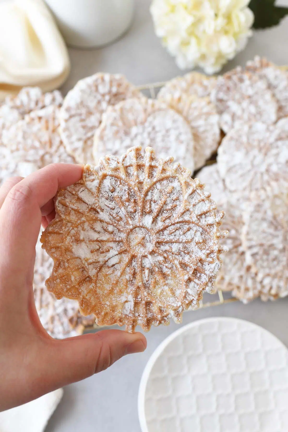 A hand holding a powdered pizzelle with more in the background.