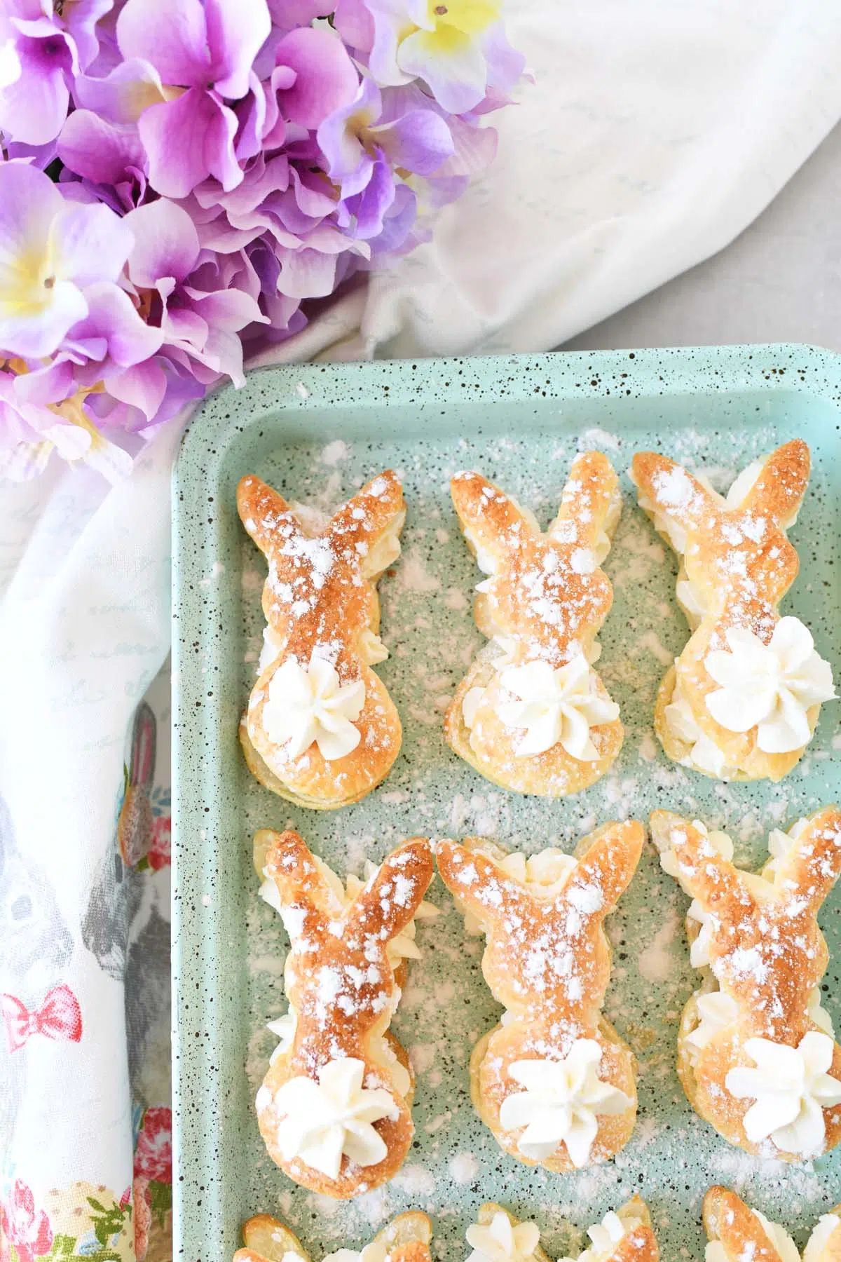 Bunny cream puffs on a blue, speckled pan near purple flowers.