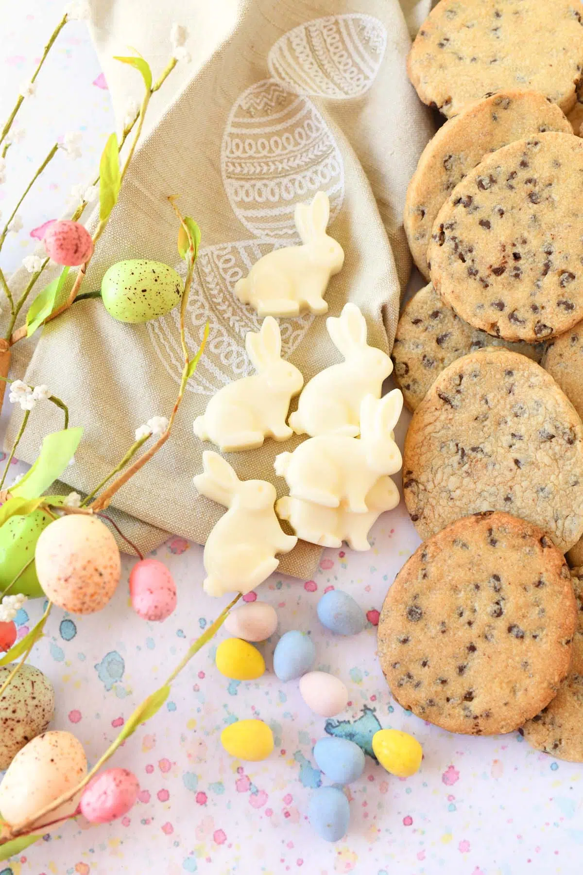 White chocolate bunnies, pastel Cadbury eggs, and chocolate chip egg cutout cookies on a pastel, speckled surface.