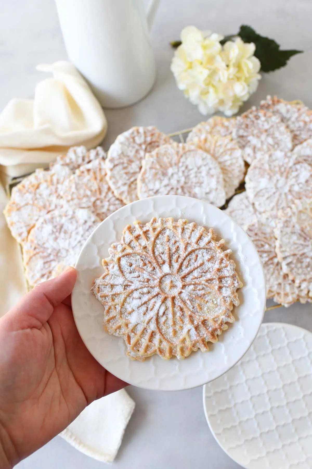A hand holding a pizzelle on a white, small plate. More pizzelles and flowers are in the background.