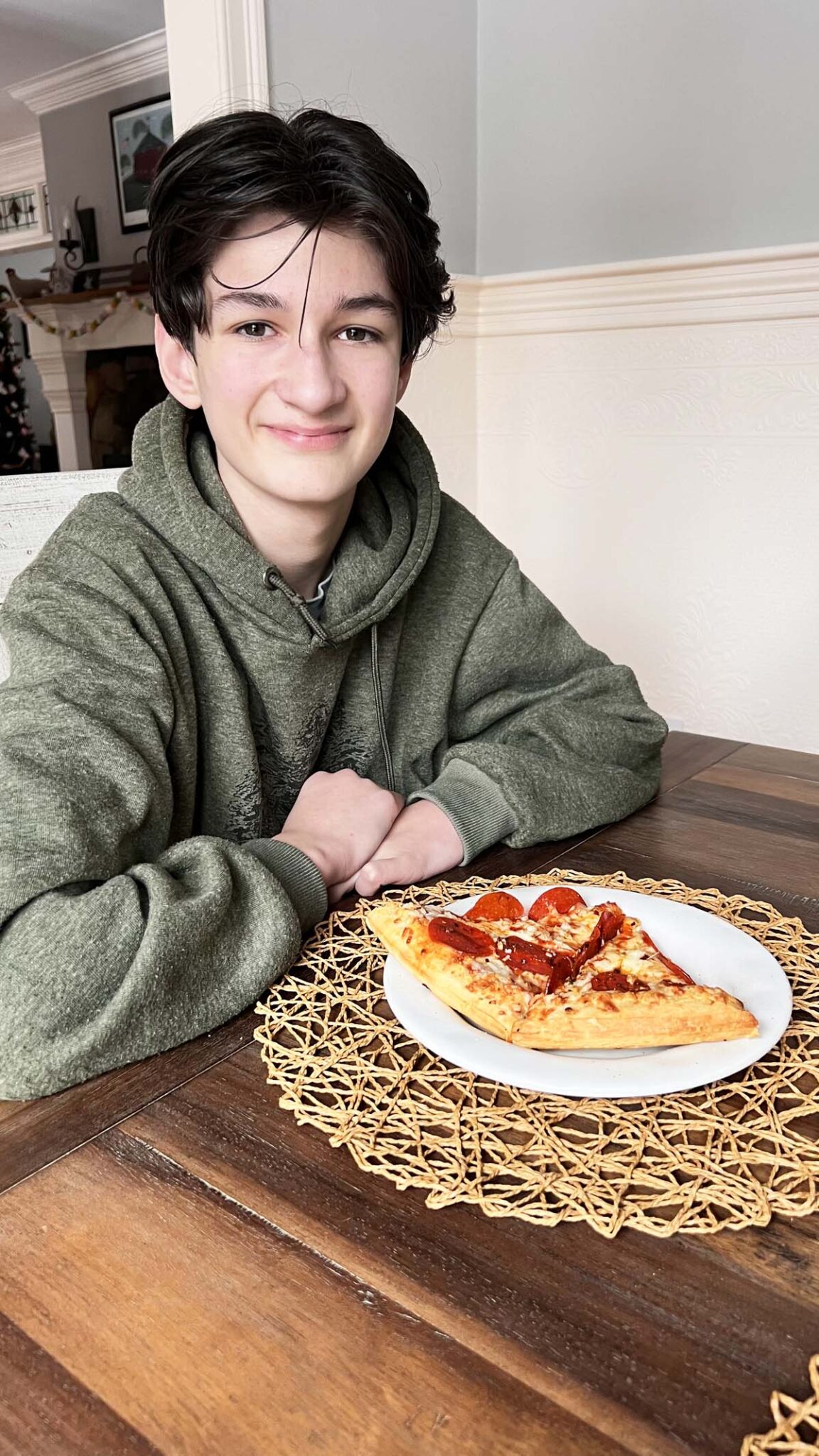 Boy in a green sweatshirt smiling with pepperoni pizza on a table.
