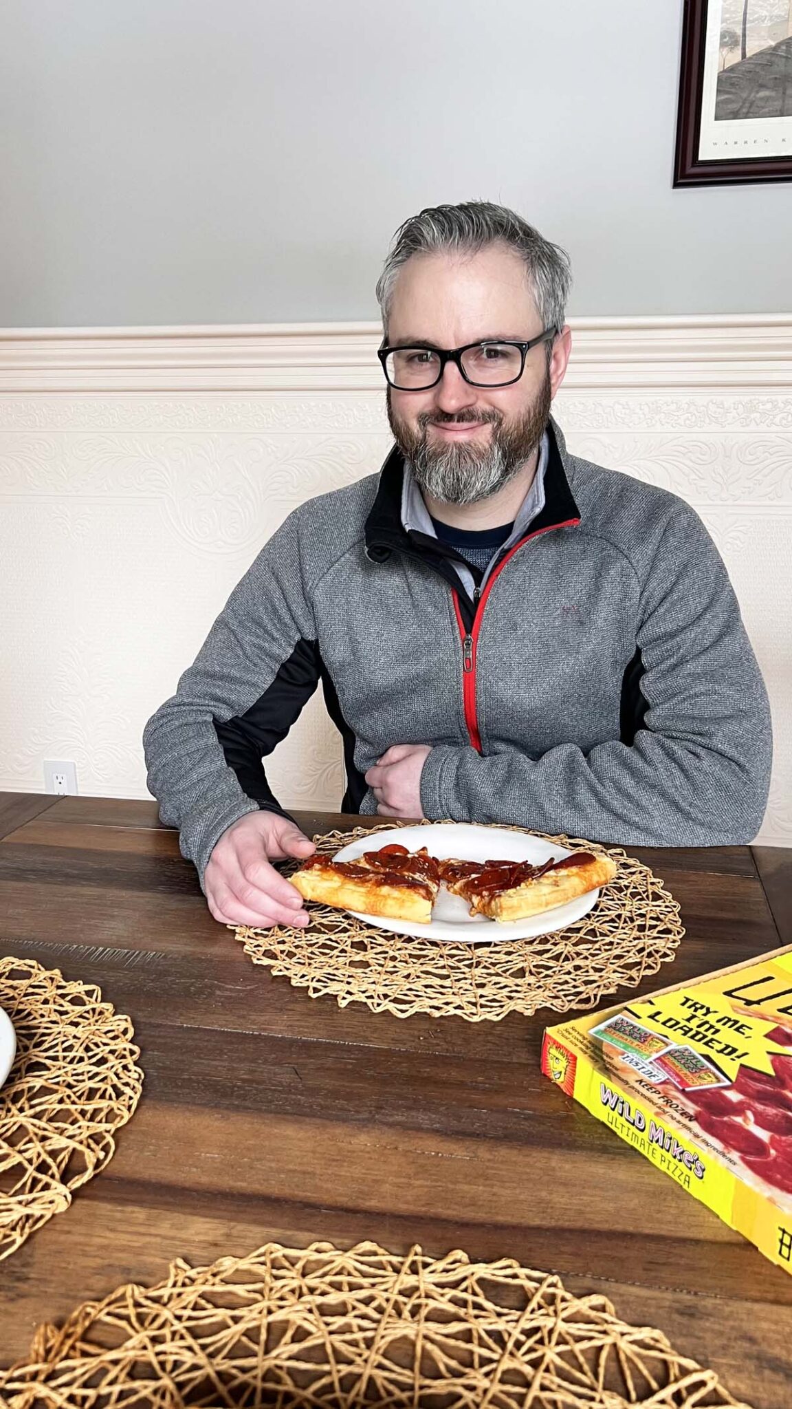 A man in grey smiling with slices of pepperoni pizza at a table.