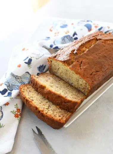Sliced sour cream banana bread on a white plate near a blue chicken napkin.