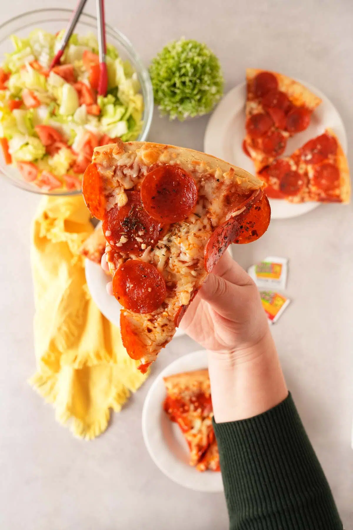 A hand holding up a slice of pepperoni pizza about a table with salad, and additional slices of pizza.
