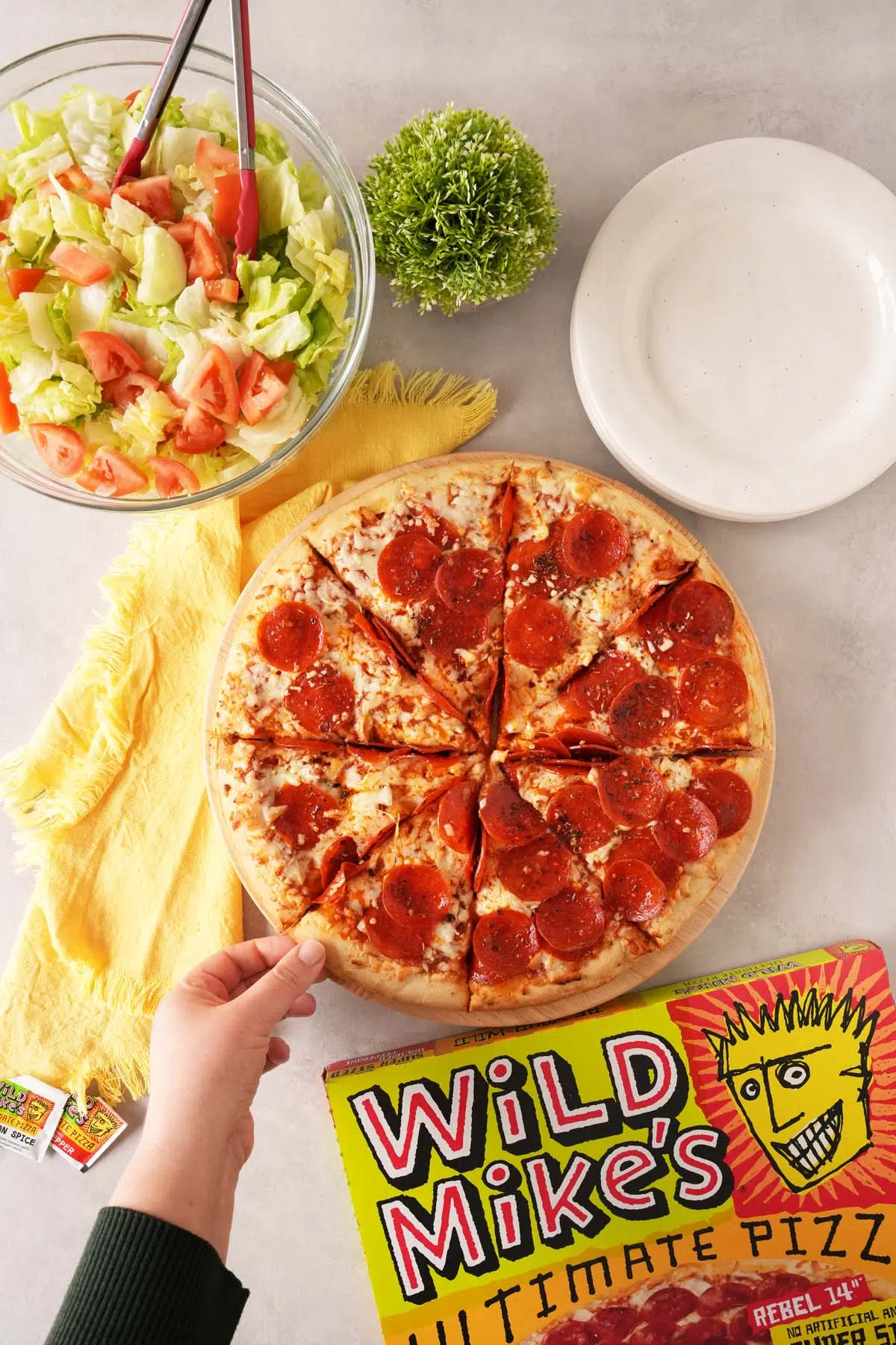 A hand grabbing a slice of Wild Mike's Pepperoni Pizza on a table setting with plates, and plant, and salad.