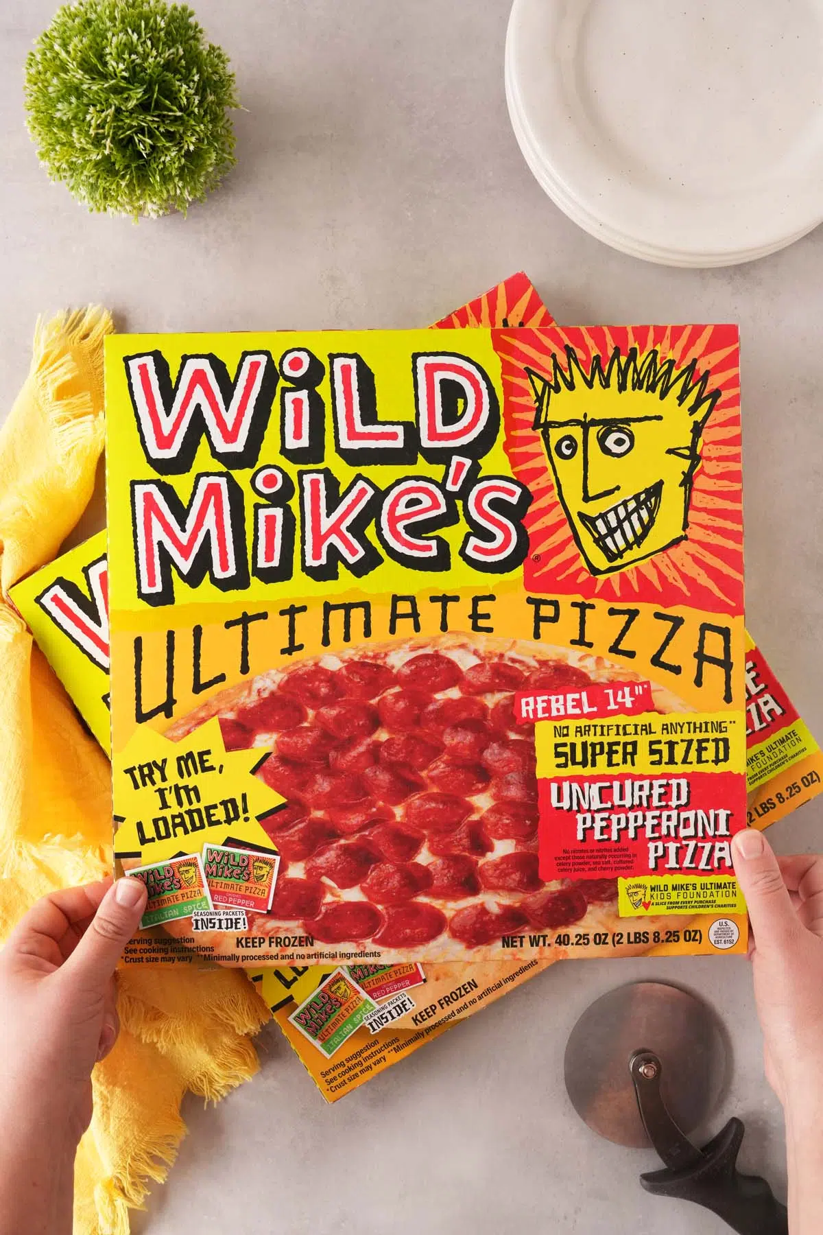 Hands holidng a box of Wild Mike's Ultimate Pizza on a grey table.