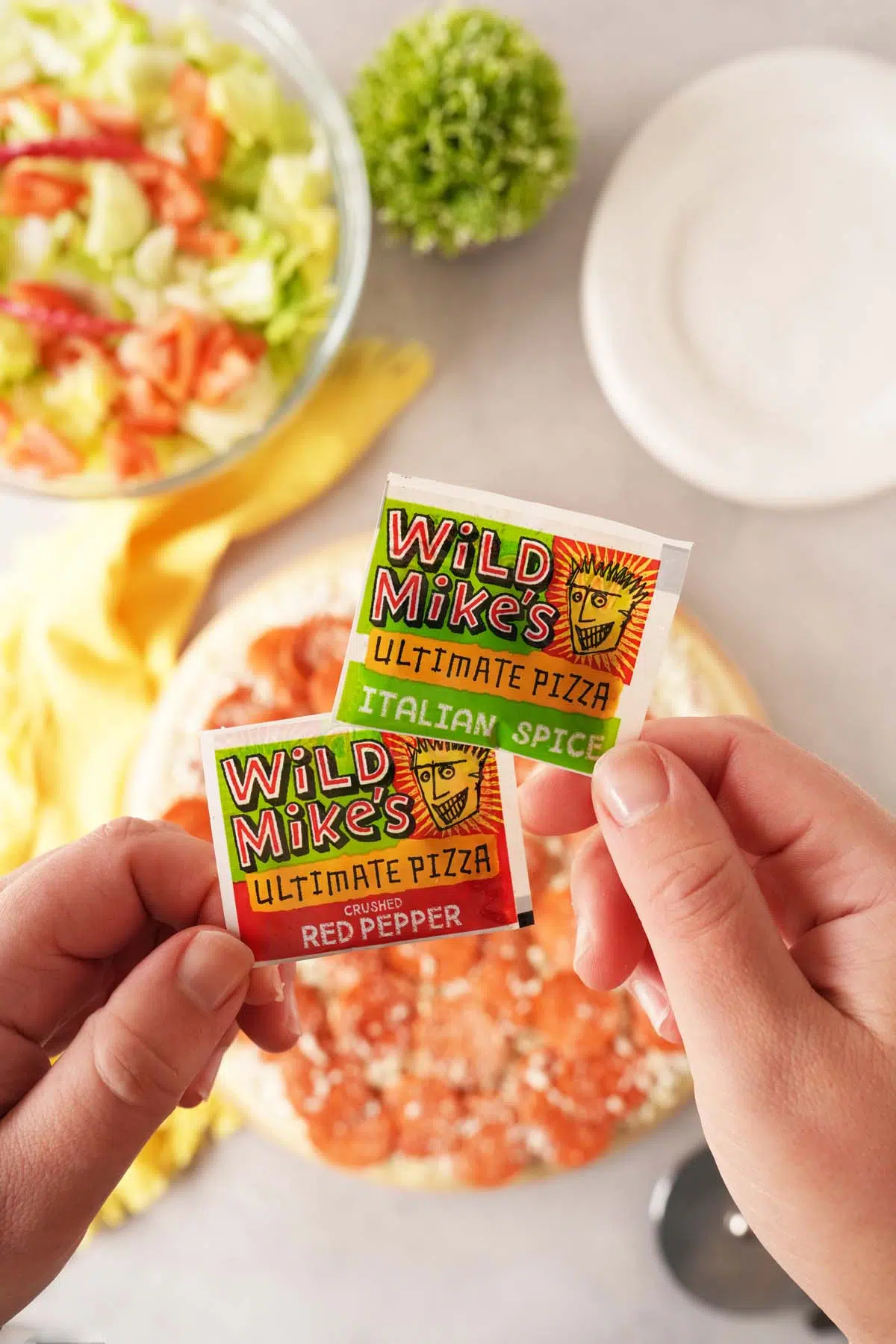 A hand holding Wild Mike's spices above a table with plates, salad, and a pizza.