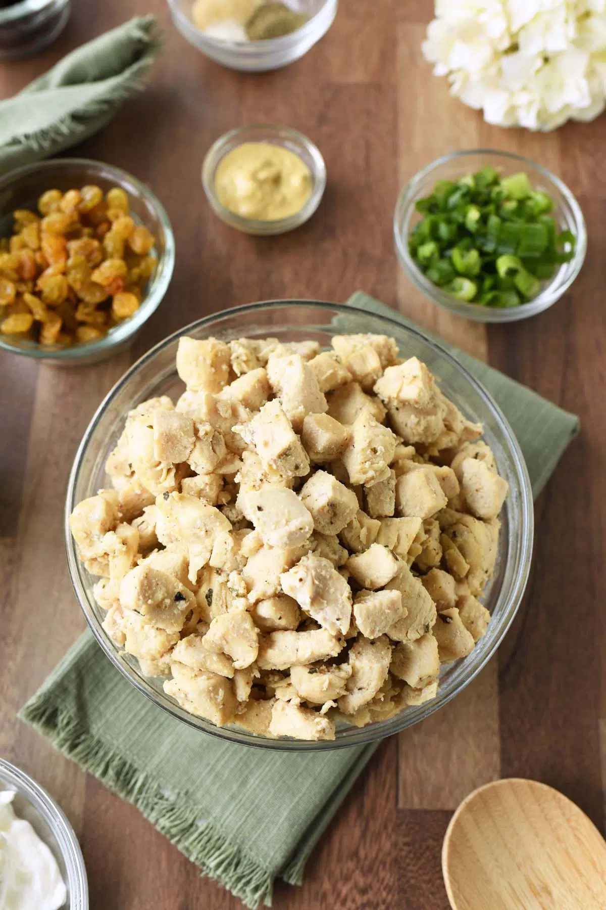 A bowl of pan-seared chicken on a wooden table with green napkins and other ingredients in small, glass bowls.