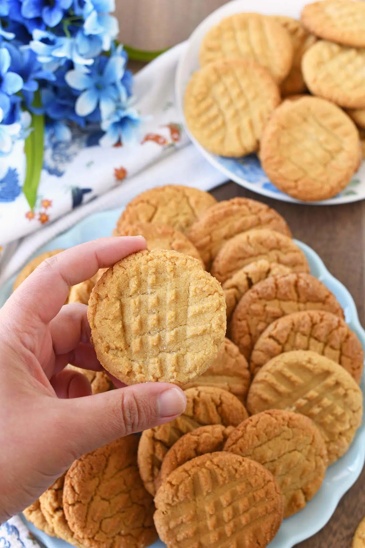 A white hand holding a peanut butter cookie near blue flowers.