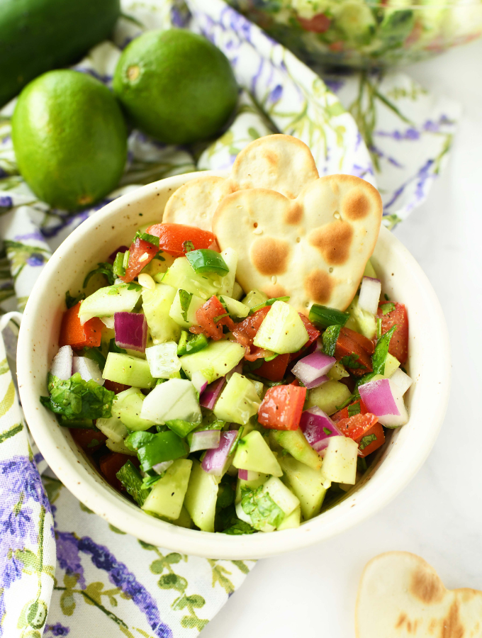 A white bowl of colorful cucumber salsa near limes and a purple, floral printed napkin.