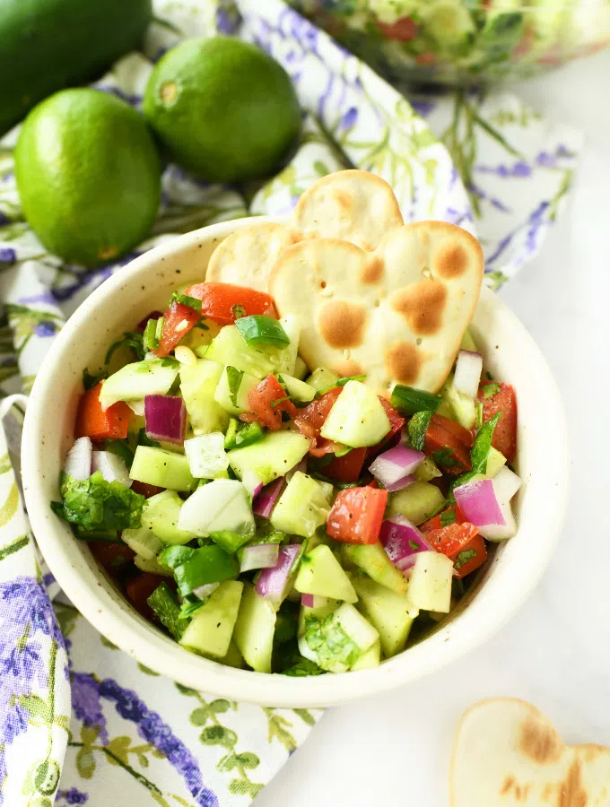 A white bowl of colorful cucumber salsa near limes and a purple, floral printed napkin.