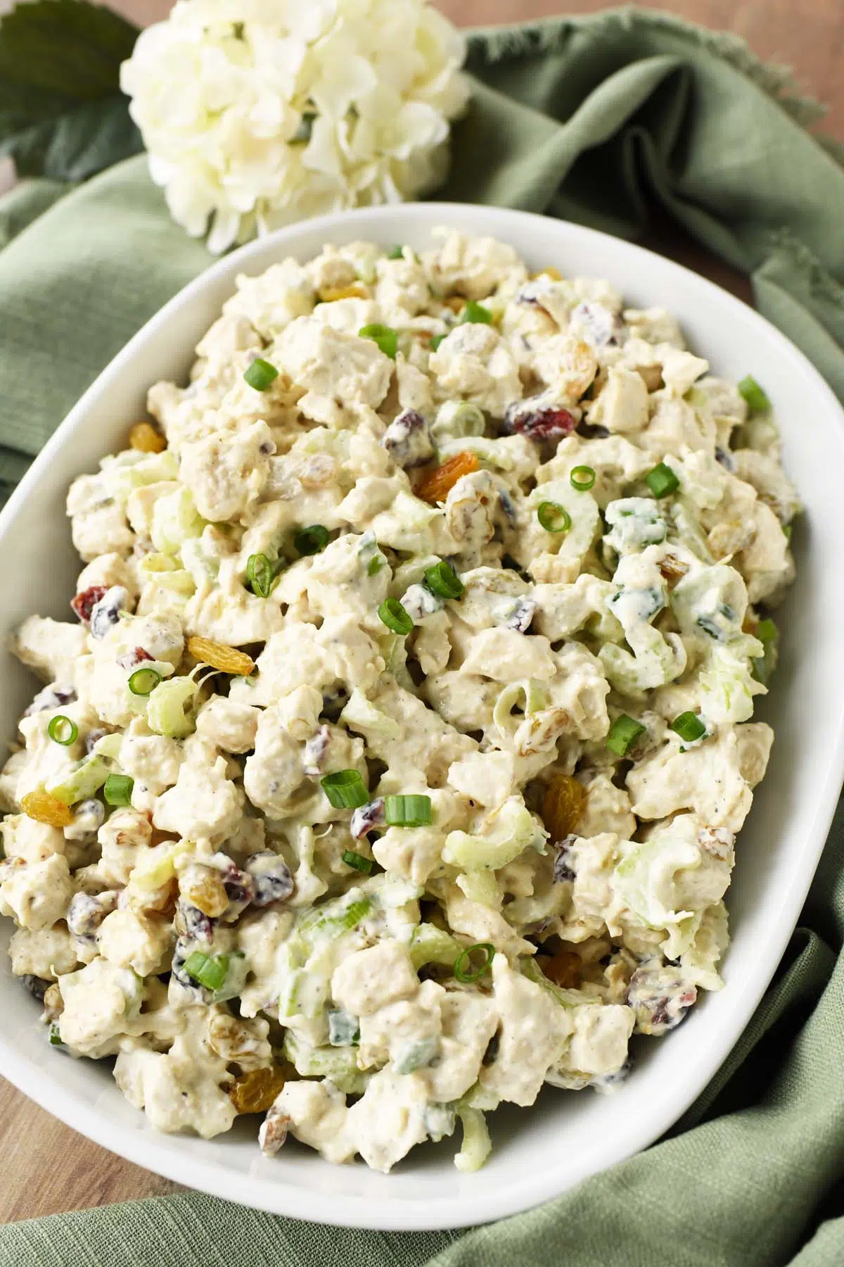 A dish of chunky chicken salad with golden raisins near a green napkin and white flower.