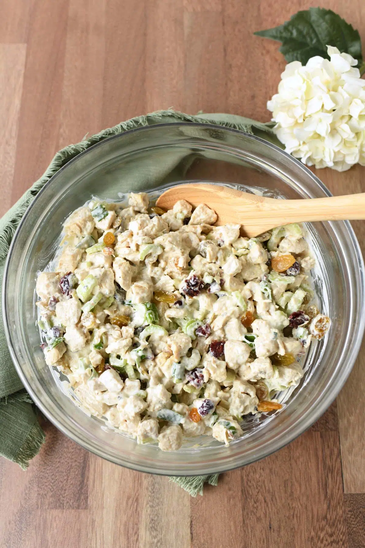 A glass bowl of harvest chicken salad with a white flower, and green napkin nearby.