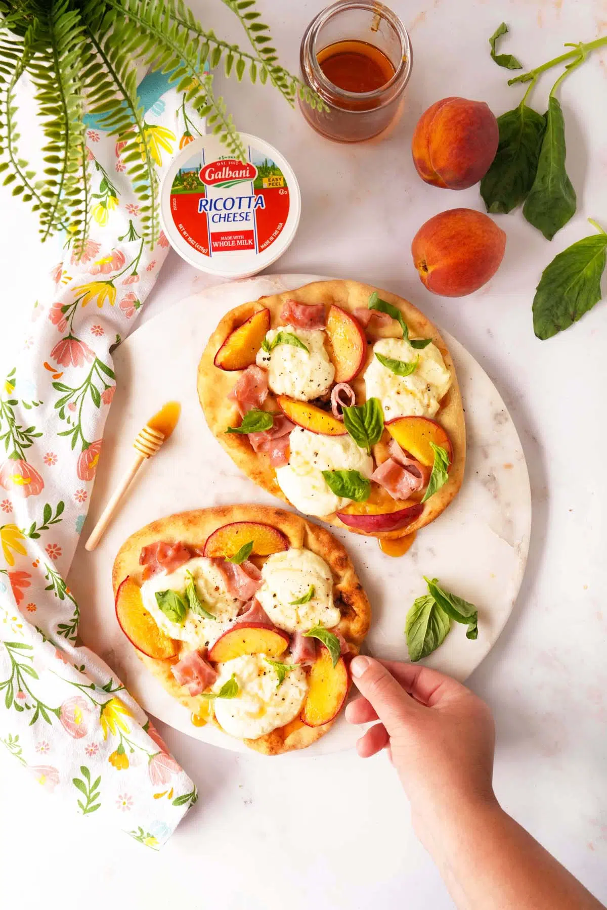 Colorful Peach Ricotta Flabread in a summery setting with a hand adding basil.