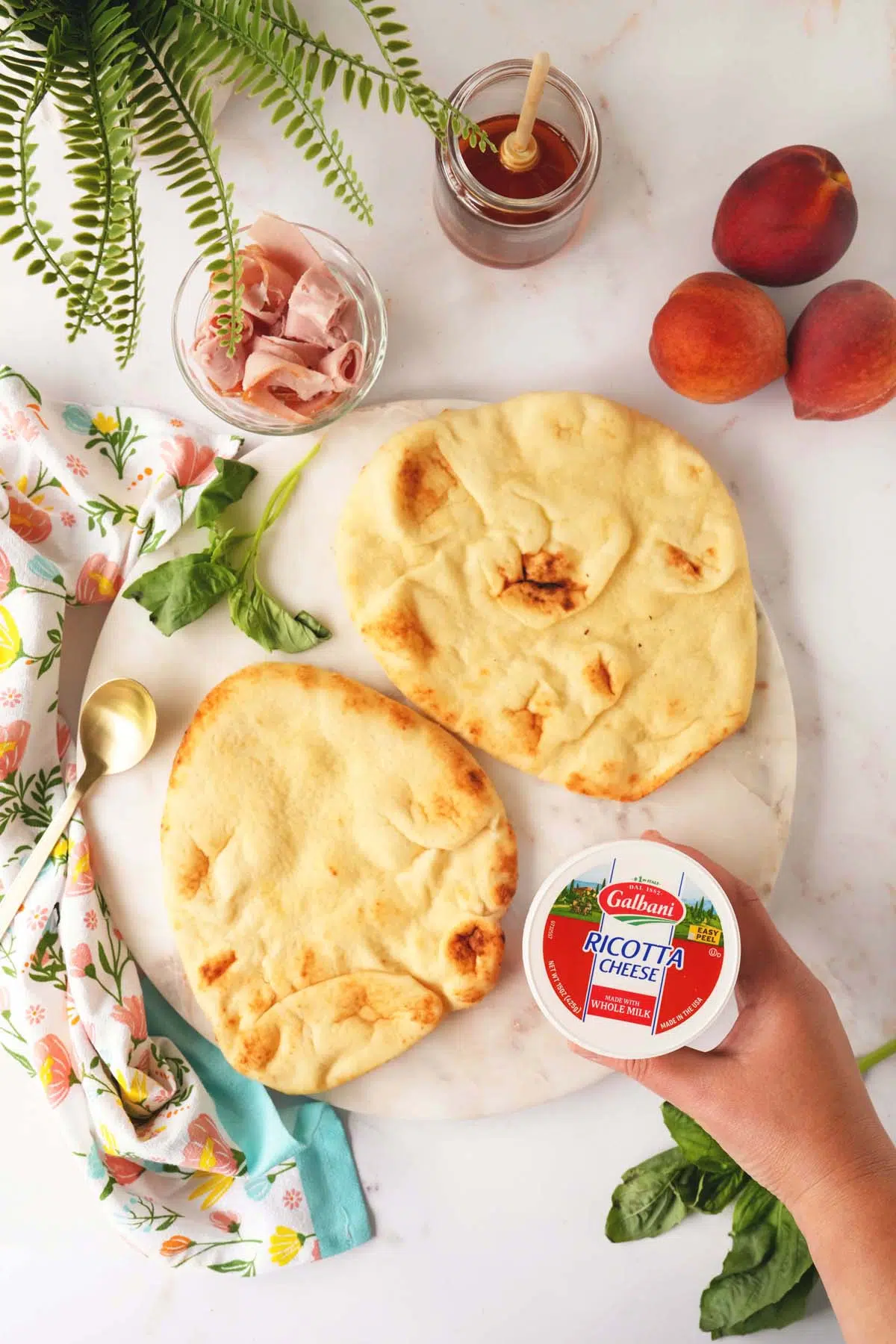 Ingredients for peach and ricotta flatbread on a white stone with a floral napkin and greens nearby.