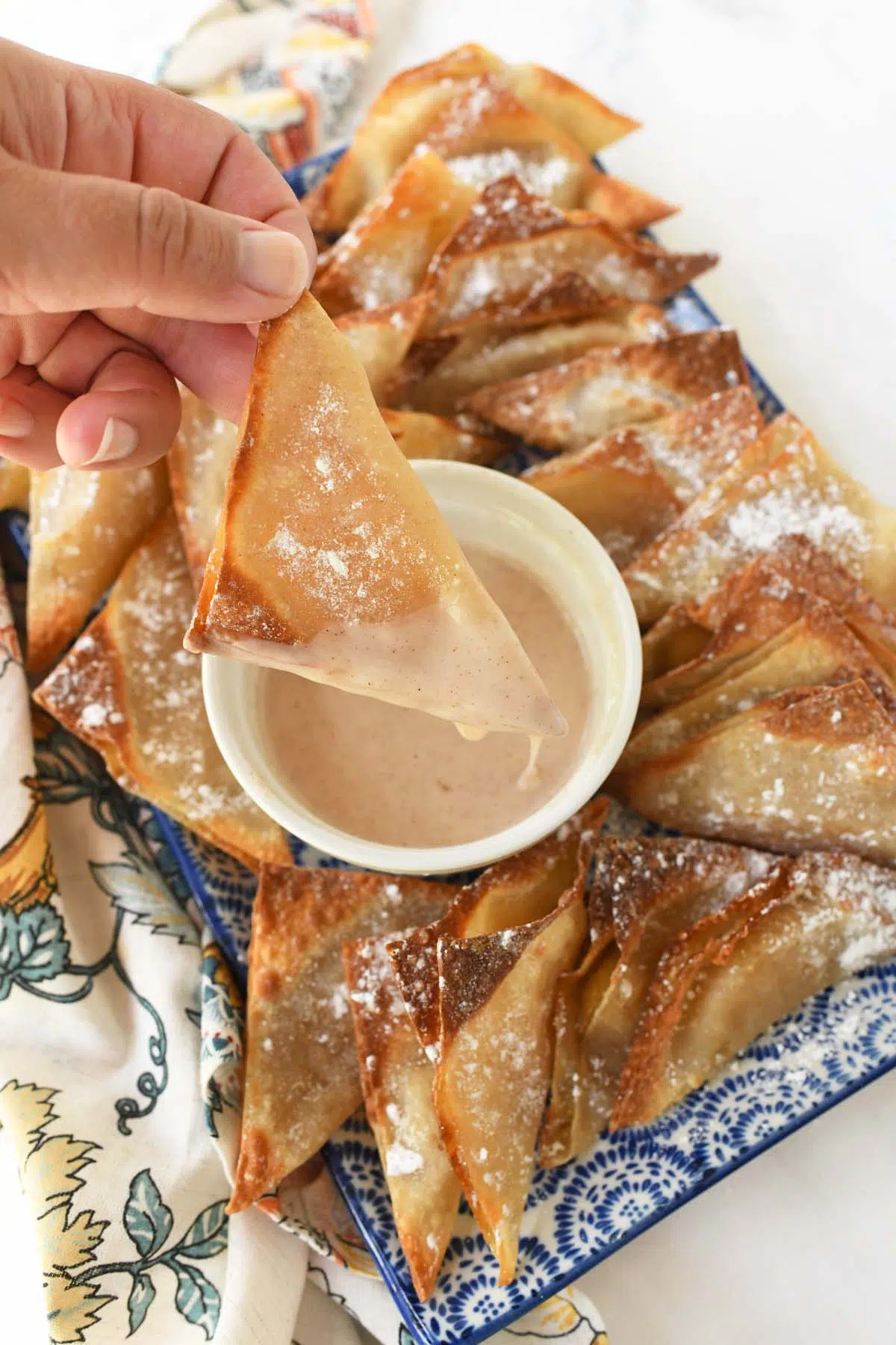 A hand holding a golden-brown apple pie wontons that is dripping with icing.