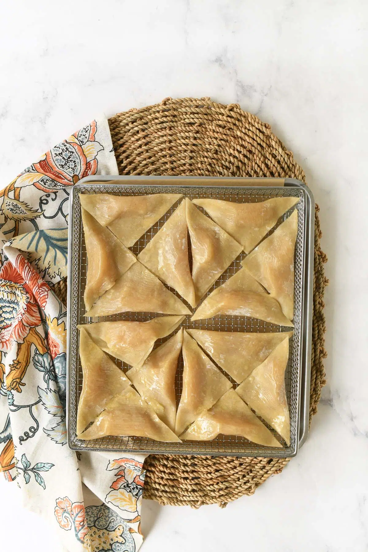 Unbaked apple pie wontons on a silver baking sheet near a patterned napkin.
