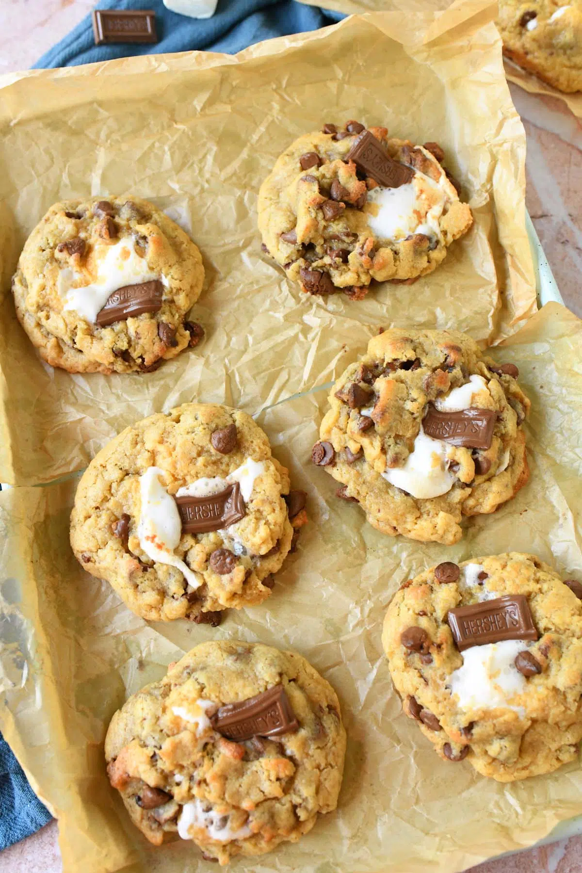 Giant baked s'mores cookies with gooey marshmallow coming out on a wrinkled parchment-lined baking sheet.