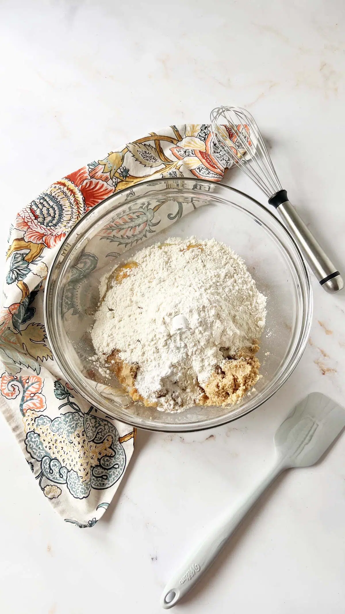 Peanut butter blondies batter in glass mixing bowl near a floral napkin, whisk, and rubber spatula.