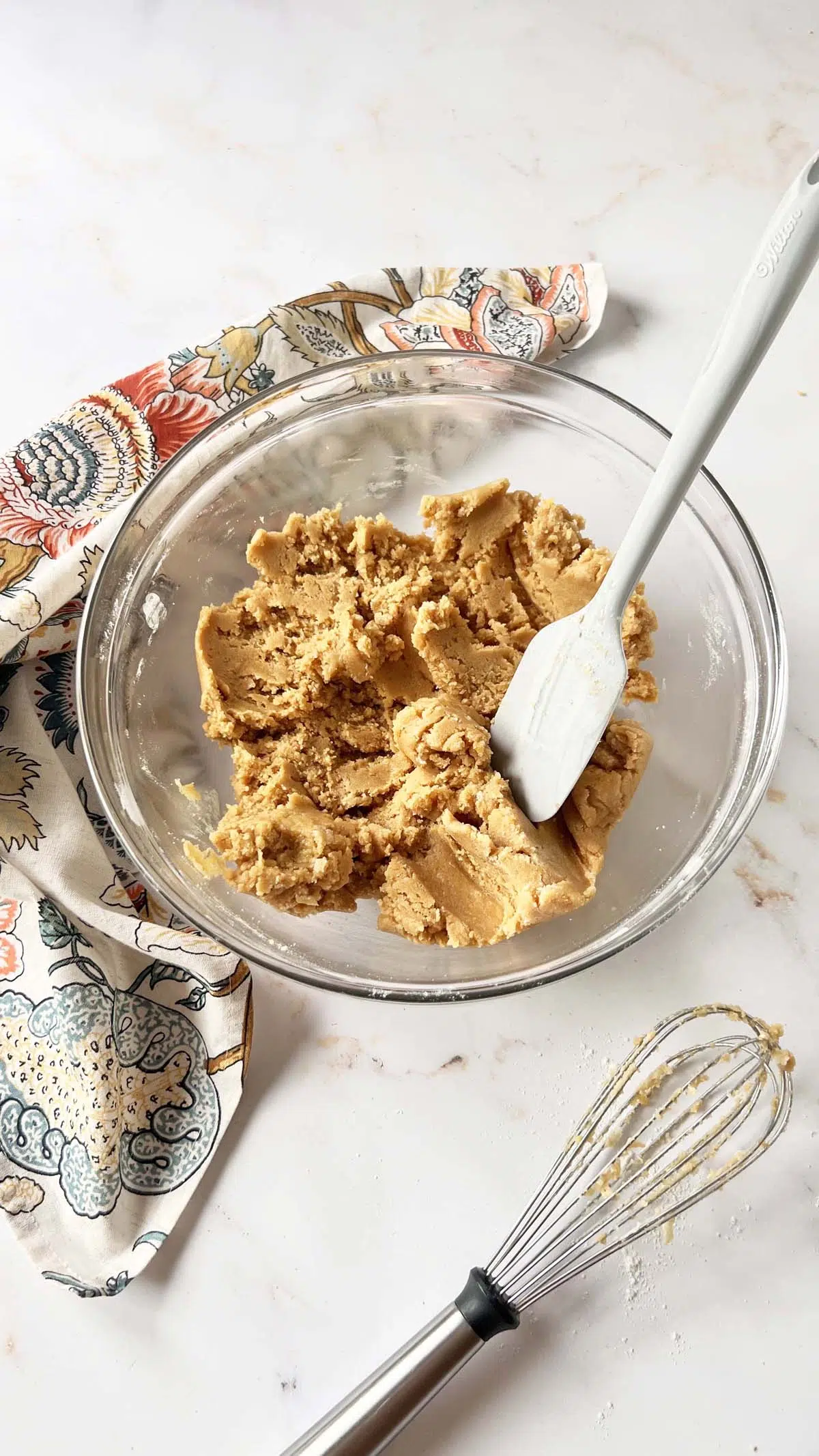 A bowl with peanut butter blondie batter near a floral napkin, rubber spatula, and whisk.