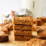 A stack on peanut butter blondies in a wooden cutting board with a white jar and floral napkin in the background.