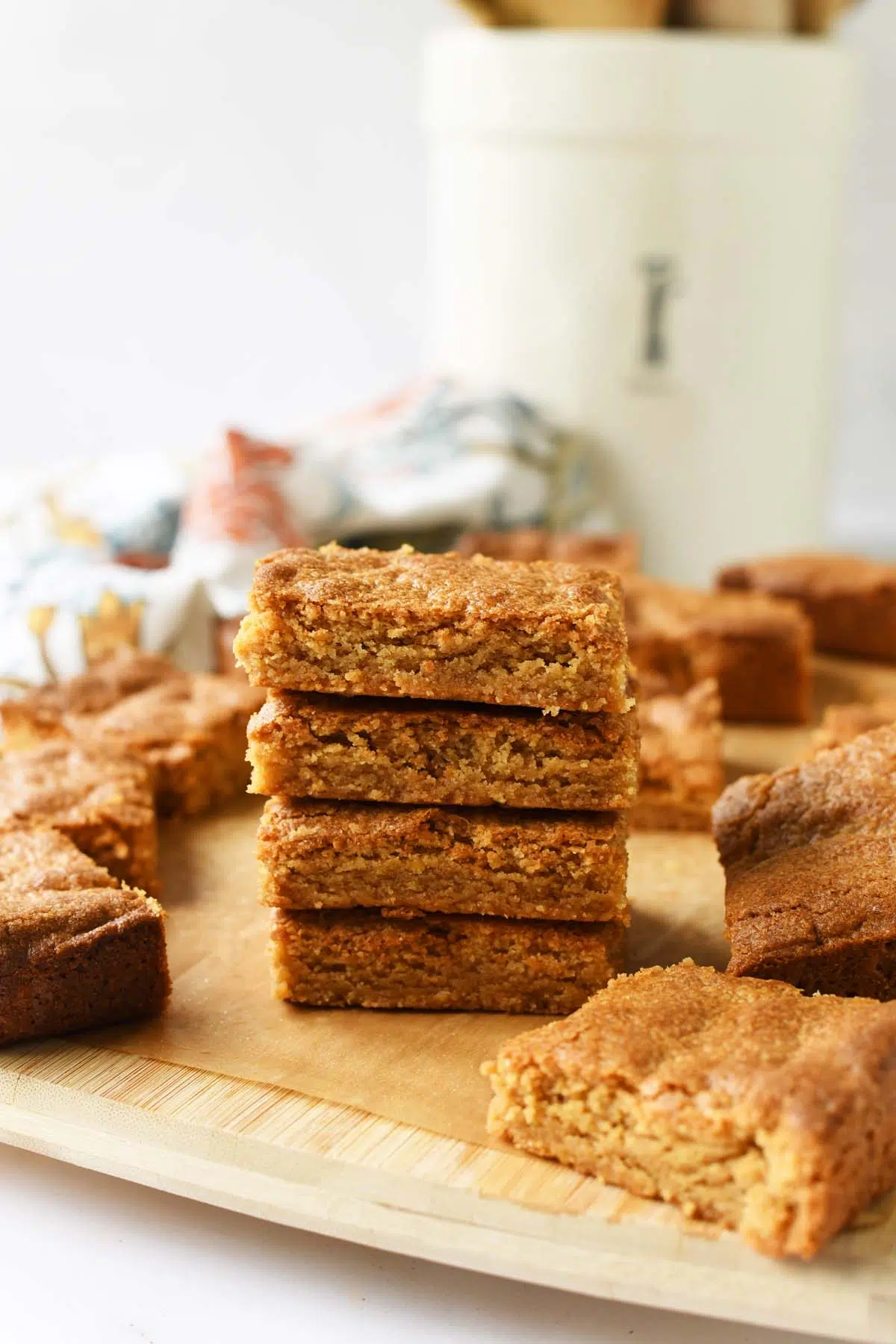 A stack on peanut butter blondies in a wooden cutting board with a white jar and floral napkin in the background.