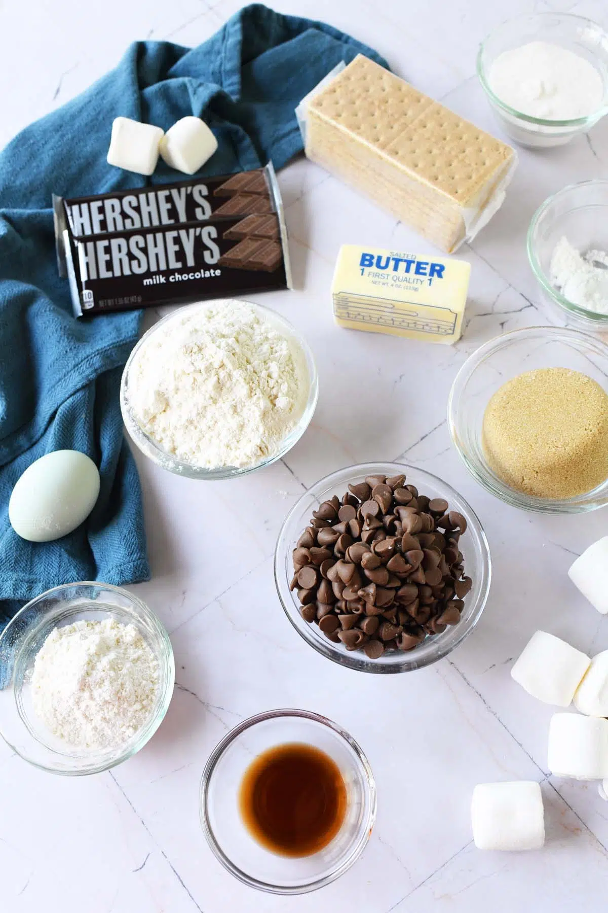 Ingredients for smores cookies in glass containers near a blue napkin on a white table.