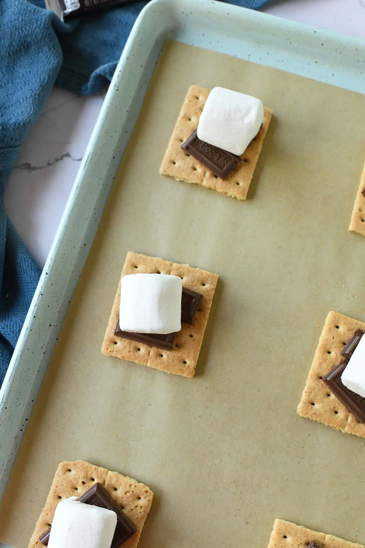 Graham crackers with chocolate and a marshmallow on a parchment-lined baking sheet near a blue napkin.