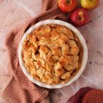 A golden-brown baked pie near a textured napkin, apples, and burgundy oven mitts.