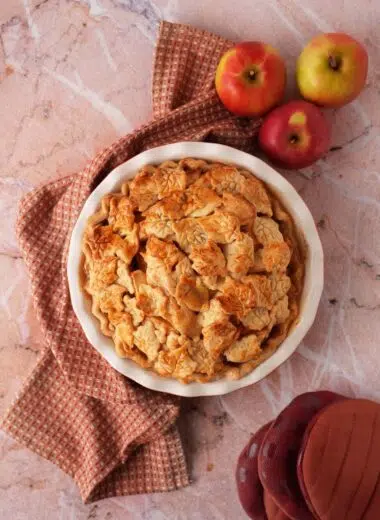 A golden-brown baked pie near a textured napkin, apples, and burgundy oven mitts.