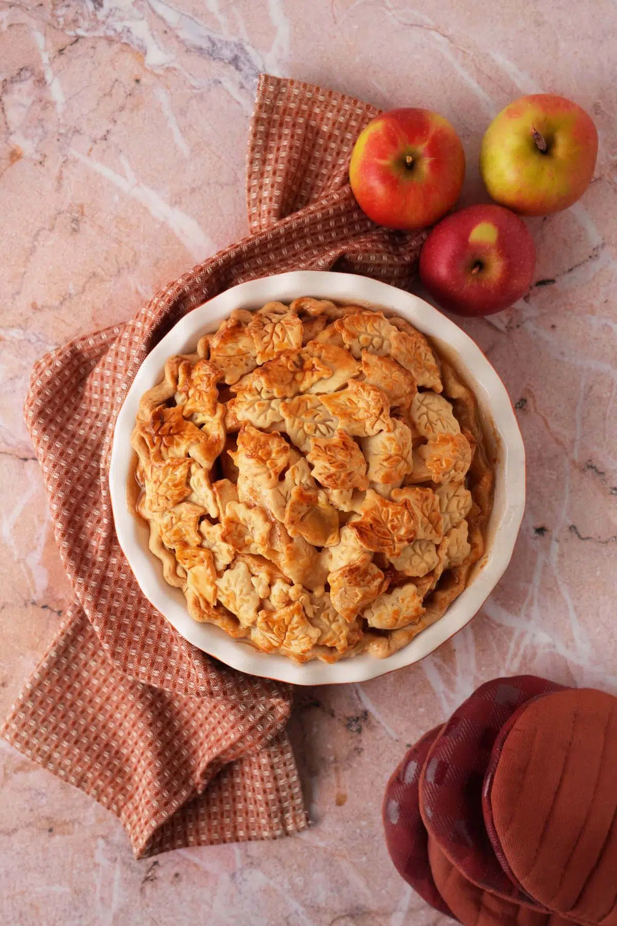 A golden-brown baked pie near a textured napkin, apples, and burgundy oven mitts.
