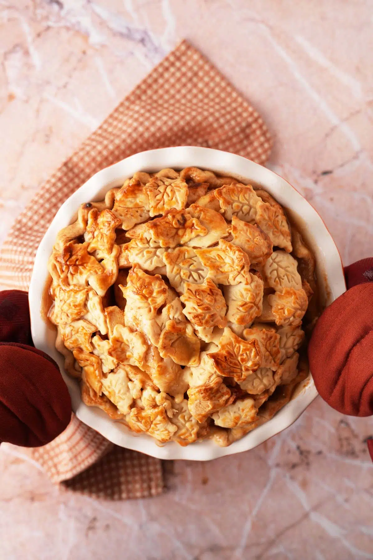 Hands in burgundy oven mitts holding up a golden-brown pie crust that is made of leaves and acorn shape pie cutouts.