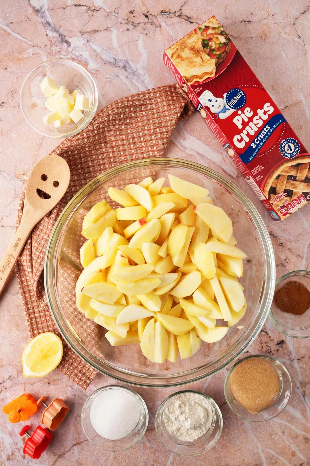 A glass bowl near apple pie ingredients, a box of pillsbury pie crust, a smiley spoon, and brown, textured napkin.