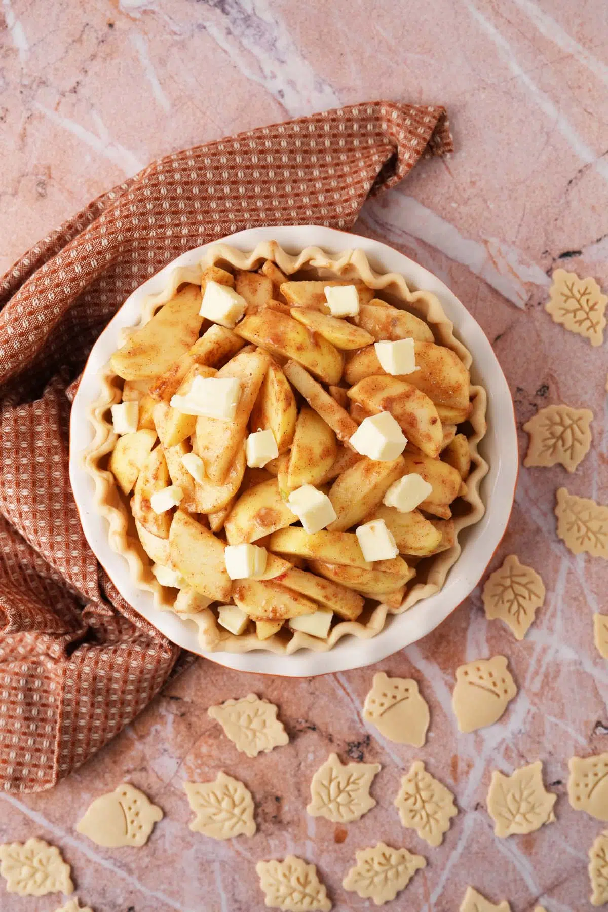 A pie that is loaded wioth seaoned apple wedges and butter pats. There is a brown, textured napkin nearby and leaf pie crust cutouts.