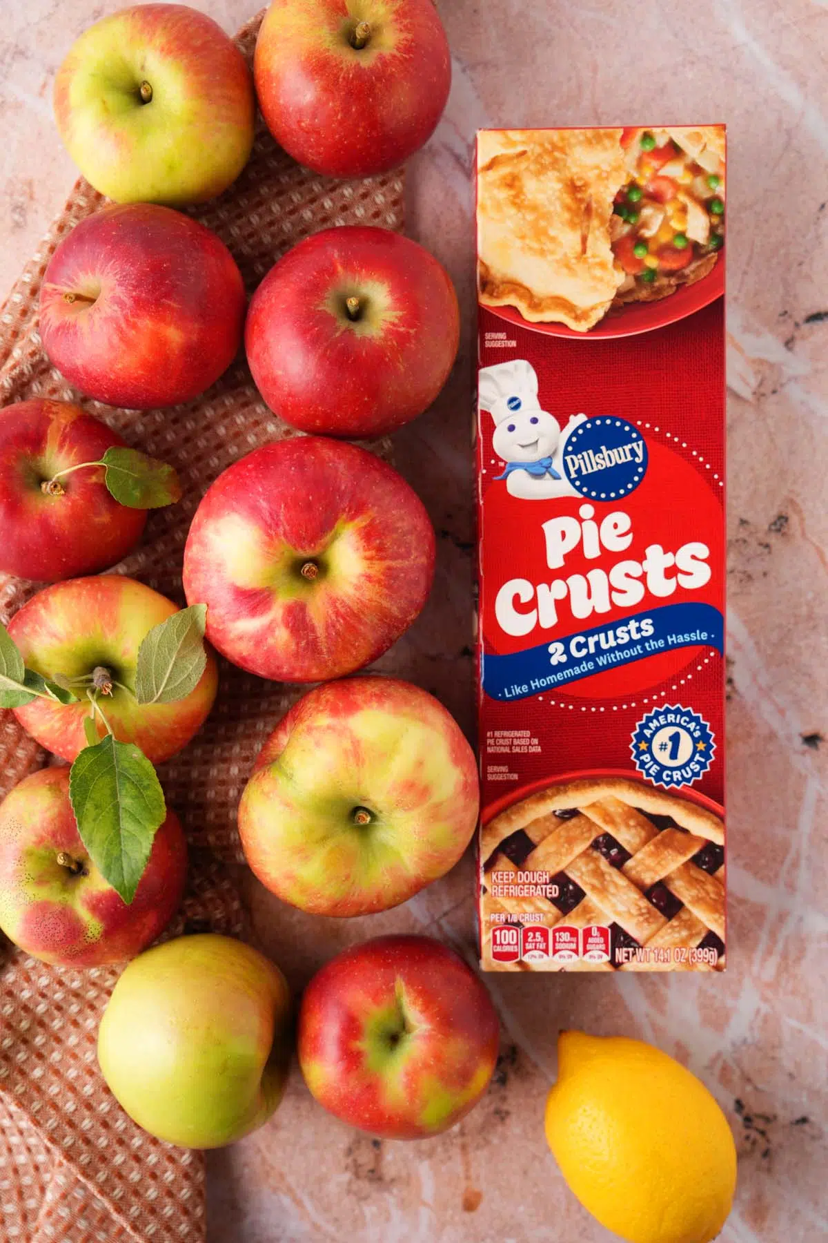 Apples that are red, yellow, and green on an earthy colored surface near a box of pillsbury pie crust, and a yellow lemon.