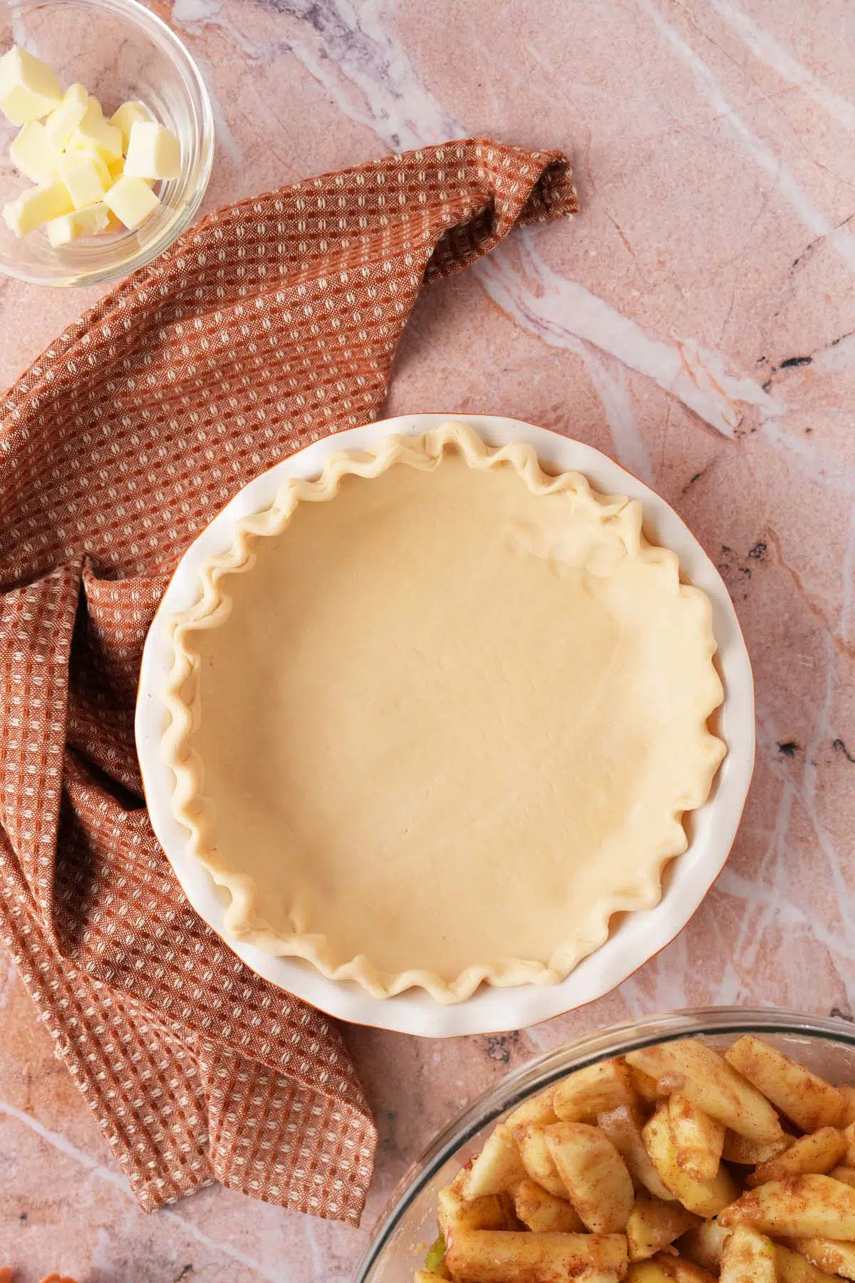 A fluted pie crust near a brown, textured napkin and a bowl of spiced apple wedges.