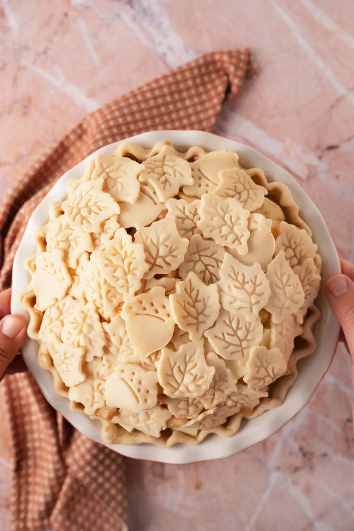 A pie covered in unbaked leaf and acorn cutouts near a brown, textured napkin.