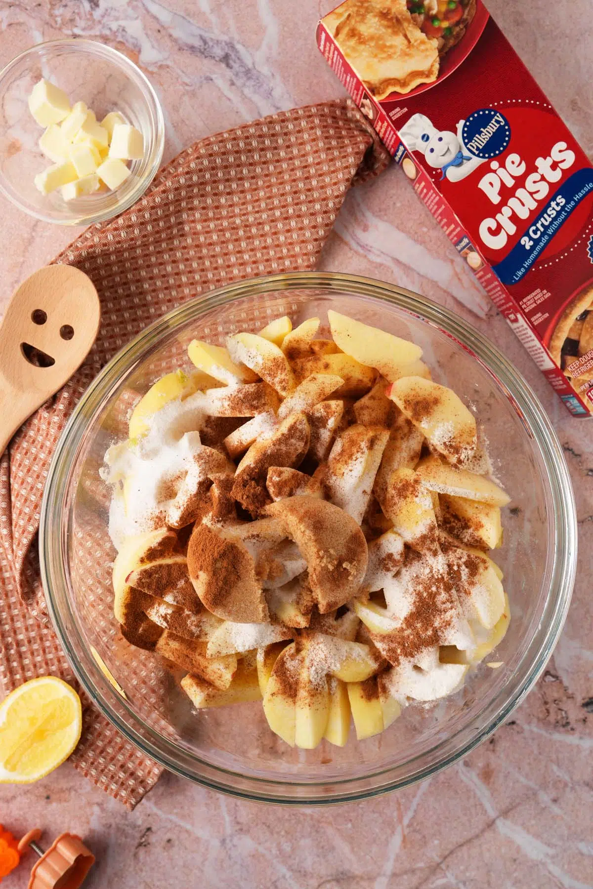 A glass bowl with apple wedges that are toppped with apple pie filling ingredients near a pie crust box and smiley spoon.